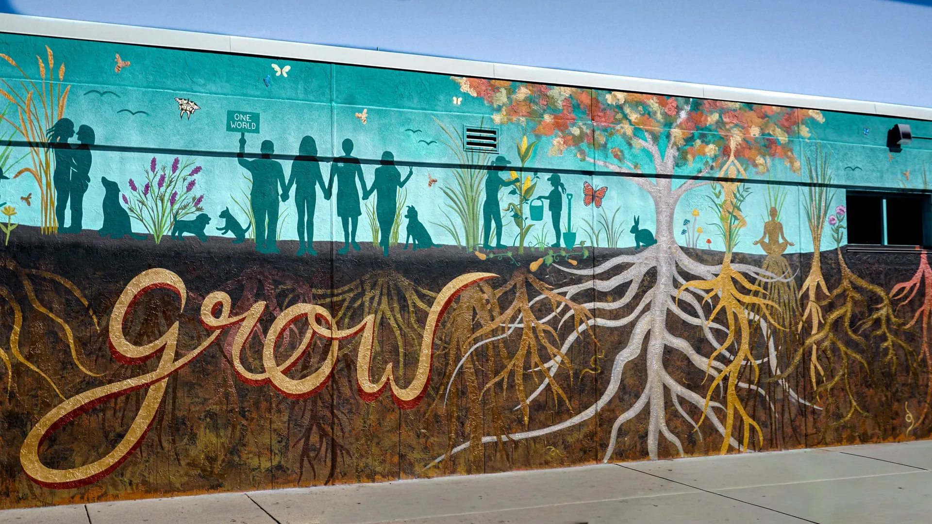 Full view of the Roots of Our Community mural: a gold 'grow' script, silhouetted neighbors holding hands, a tree, and white and gold root systems above and below ground.